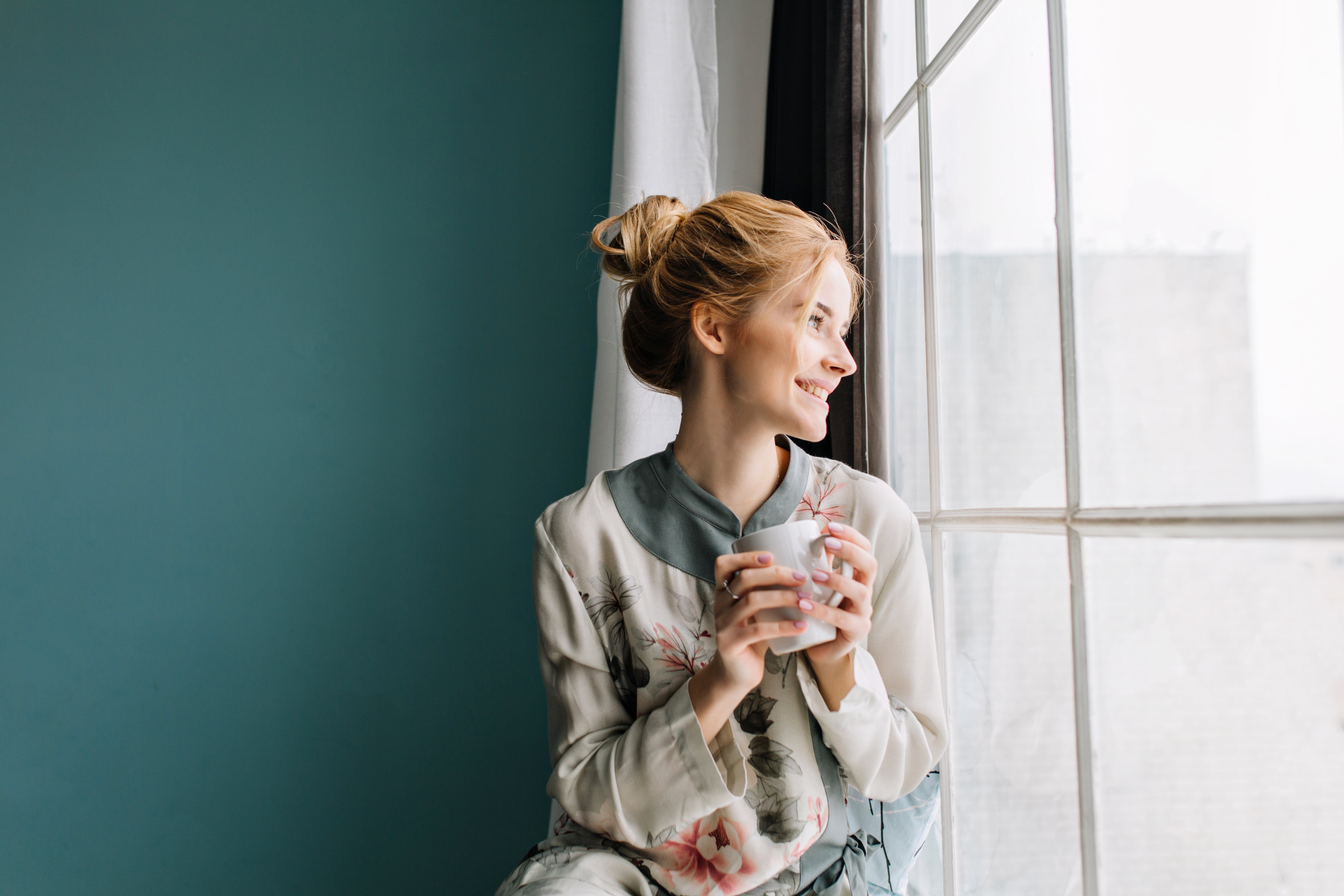 portrait-young-woman-with-blonde-hair-drinking-coffee-tea-big-window-smiling-enjoying-happy-morning-home-turquoise-wall-wearing-silk-pajamas-flowers.jpg